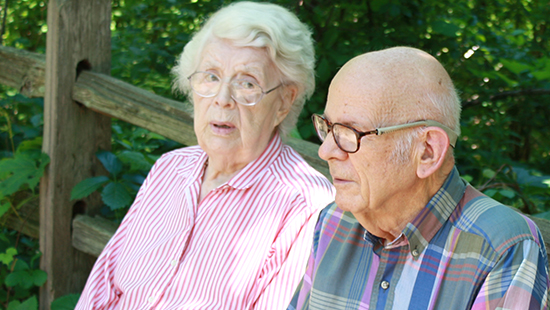 An older couple sitting outdoors on a wooden bench, surrounded by greenery
